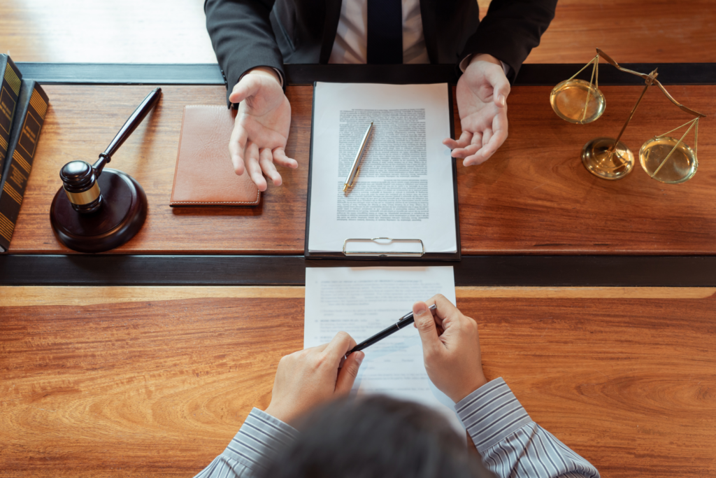 Two professionals discuss a contract at a wooden desk, with a gavel, clipboard, and scales nearby.